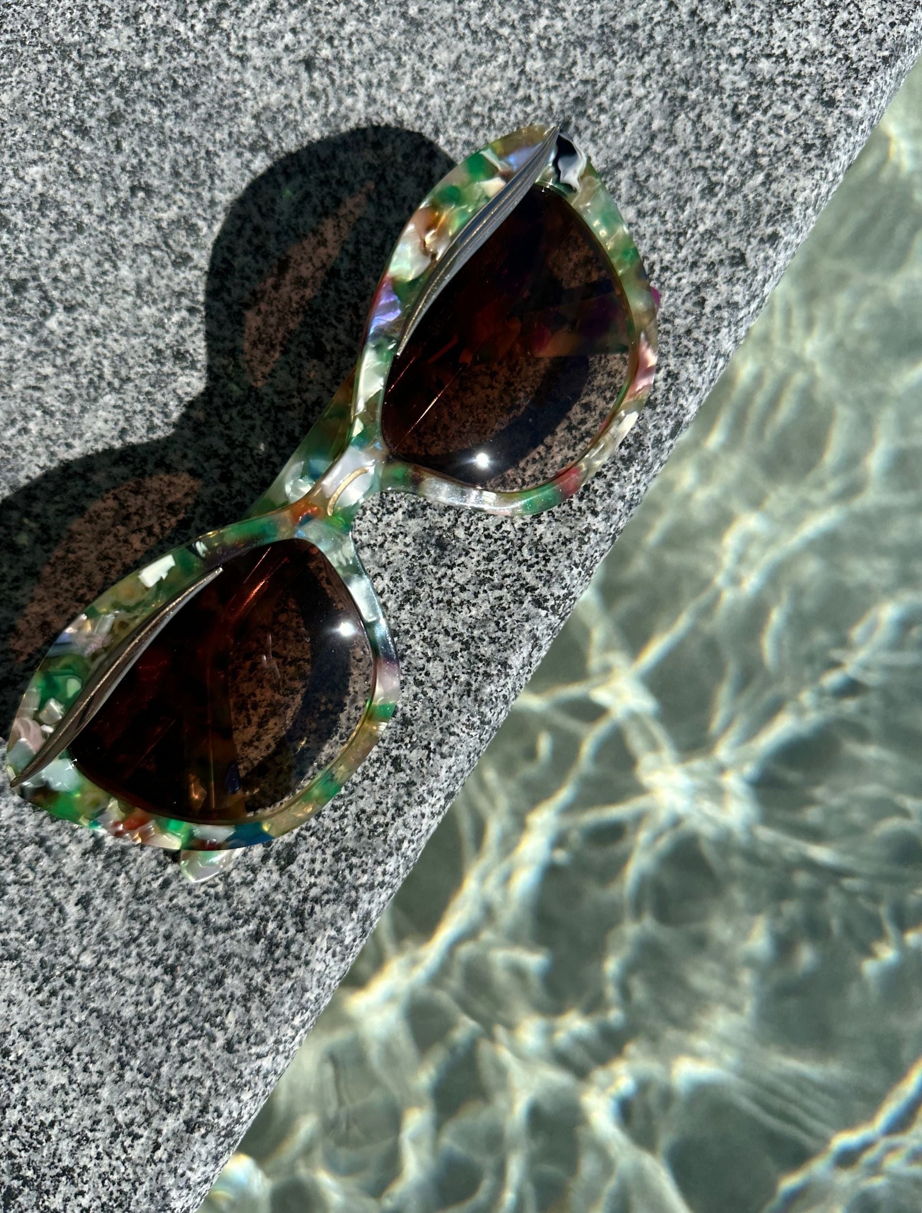 Multi-coloured cat eye sunglasses with a mix of green, orange and pearl tones, resting on a textured stone surface near shimmering water, reflecting light patterns