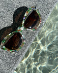 Multi-coloured cat eye sunglasses with a mix of green, orange and pearl tones, resting on a textured stone surface near shimmering water, reflecting light patterns