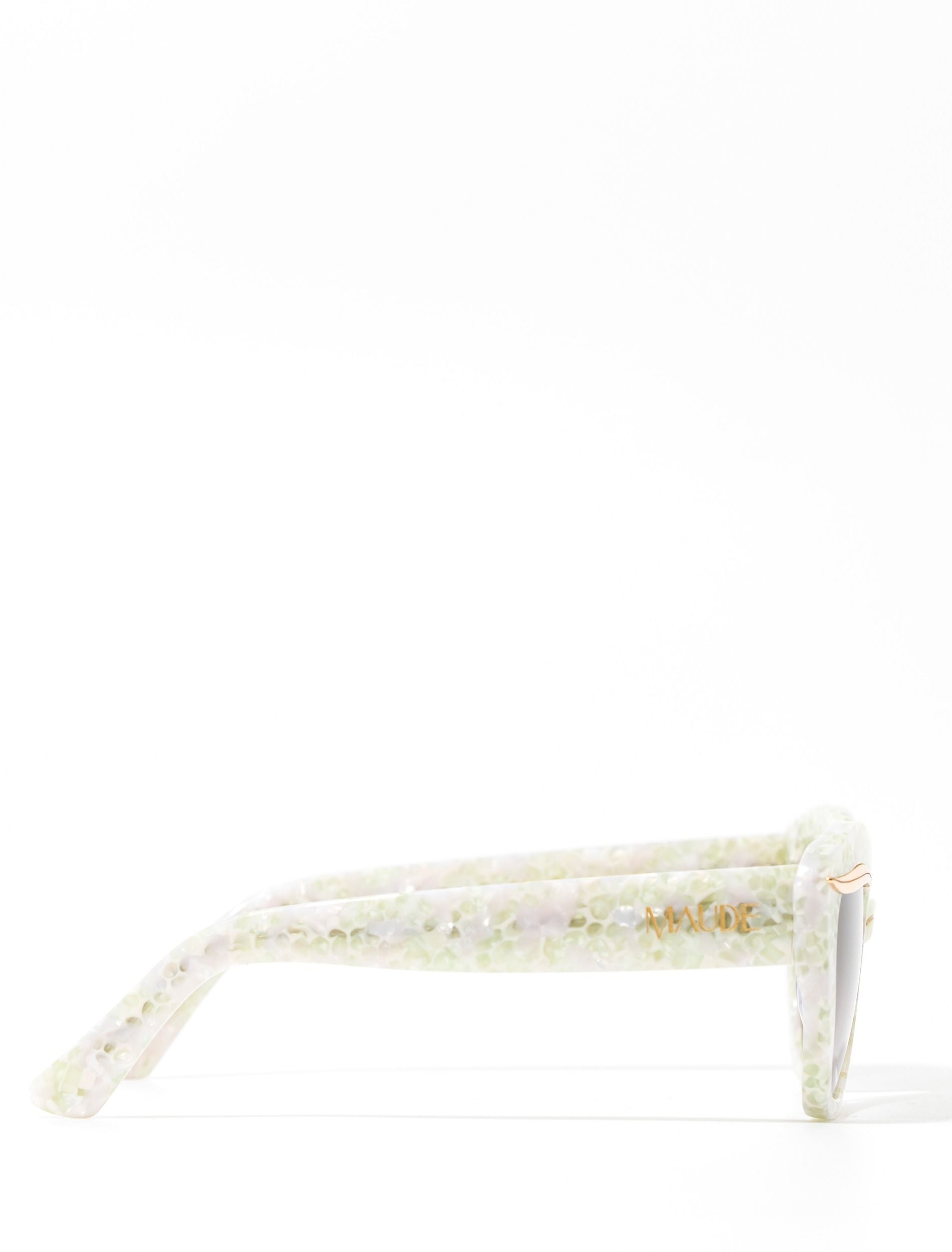 Side view of a stylish pair of sunglasses with a subtle white pearl frame, set against a white backdrop. The words MAUDE are shown on the arm of the sunglasses
