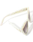 Top view of a stylish pair of sunglasses adorned with a gold flick along the upper lense and a subtle white pearl frame, set against a white backdrop. Featuring a dark grey gradient lense. The words MAUDE are shown on the arm of the sunglasses
