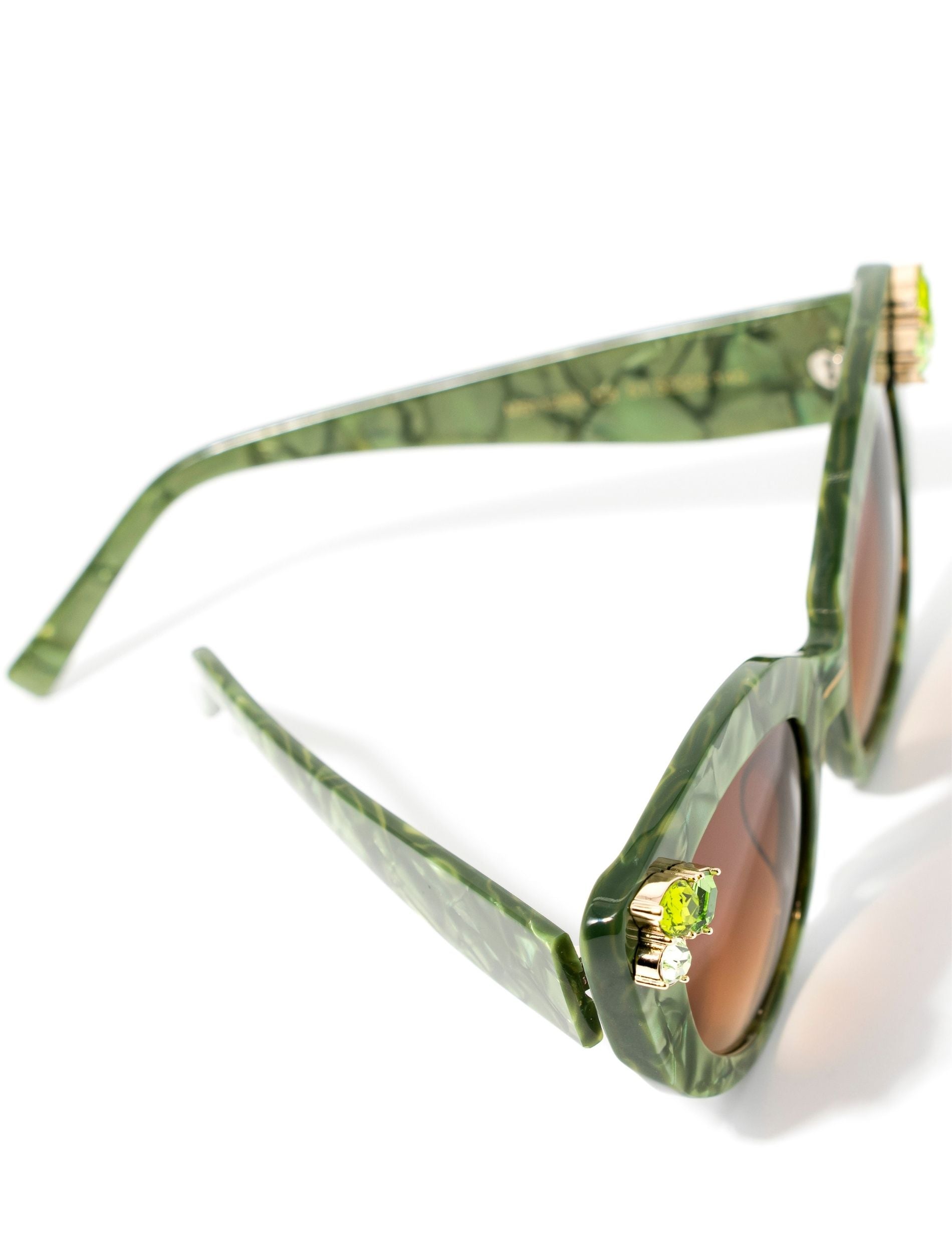 Top view of stunning cat eye sunglasses crafted with a green pearlescent look, featuring gemstone embellishments on the upper corners of the frame and a brown gradient lense. Set against a white backdrop