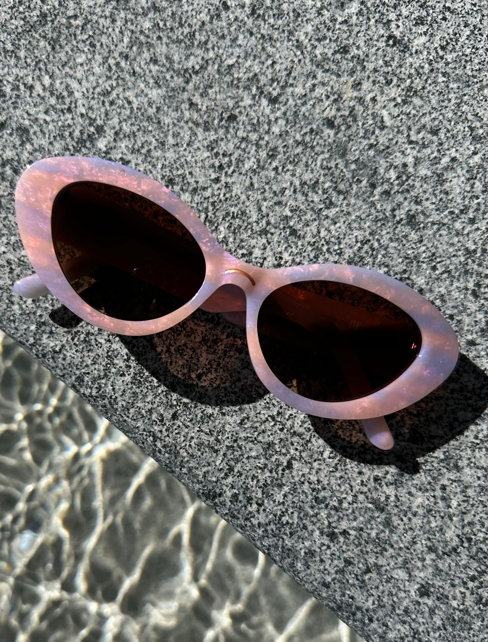 Top view of a pair of stylish pastel pink and lilac sunglasses, resting on a textured stone, highlighting their reflective frames and modern style