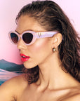 A model with brown hair and glossy red lips wears a pair of pastel pink and purple framed sunglasses in front of a pink sunset background