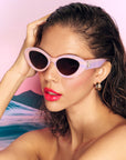 A model with brown hair and glossy red lips wears a pair of pastel pink and purple framed sunglasses in front of a pink sunset background