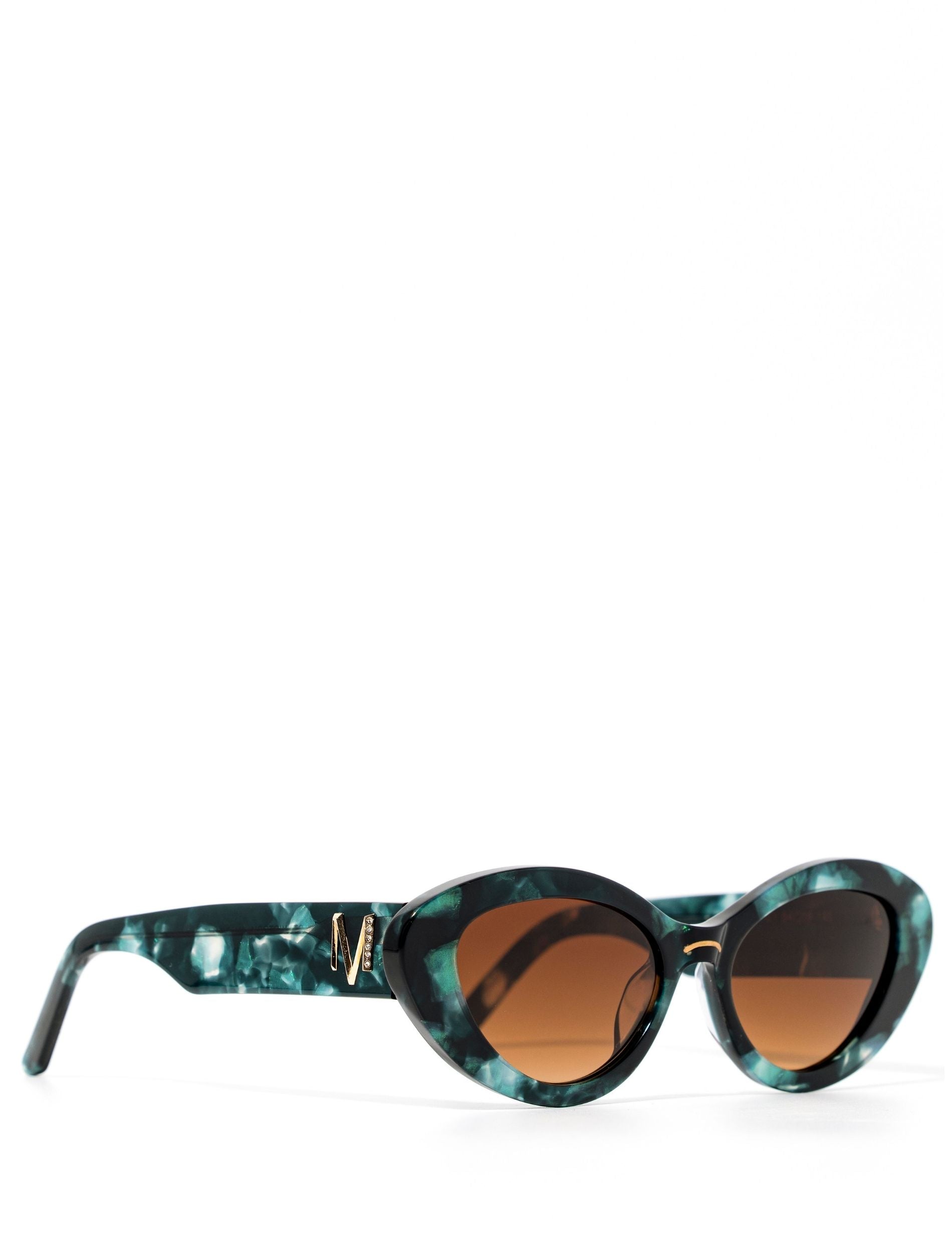 side view of a pair of stylish sunglasses in a green tortoise shell style displaced against a white back drop. the lenses are a gradient brown. You can see an embellished 'M' on the side of the frame arm