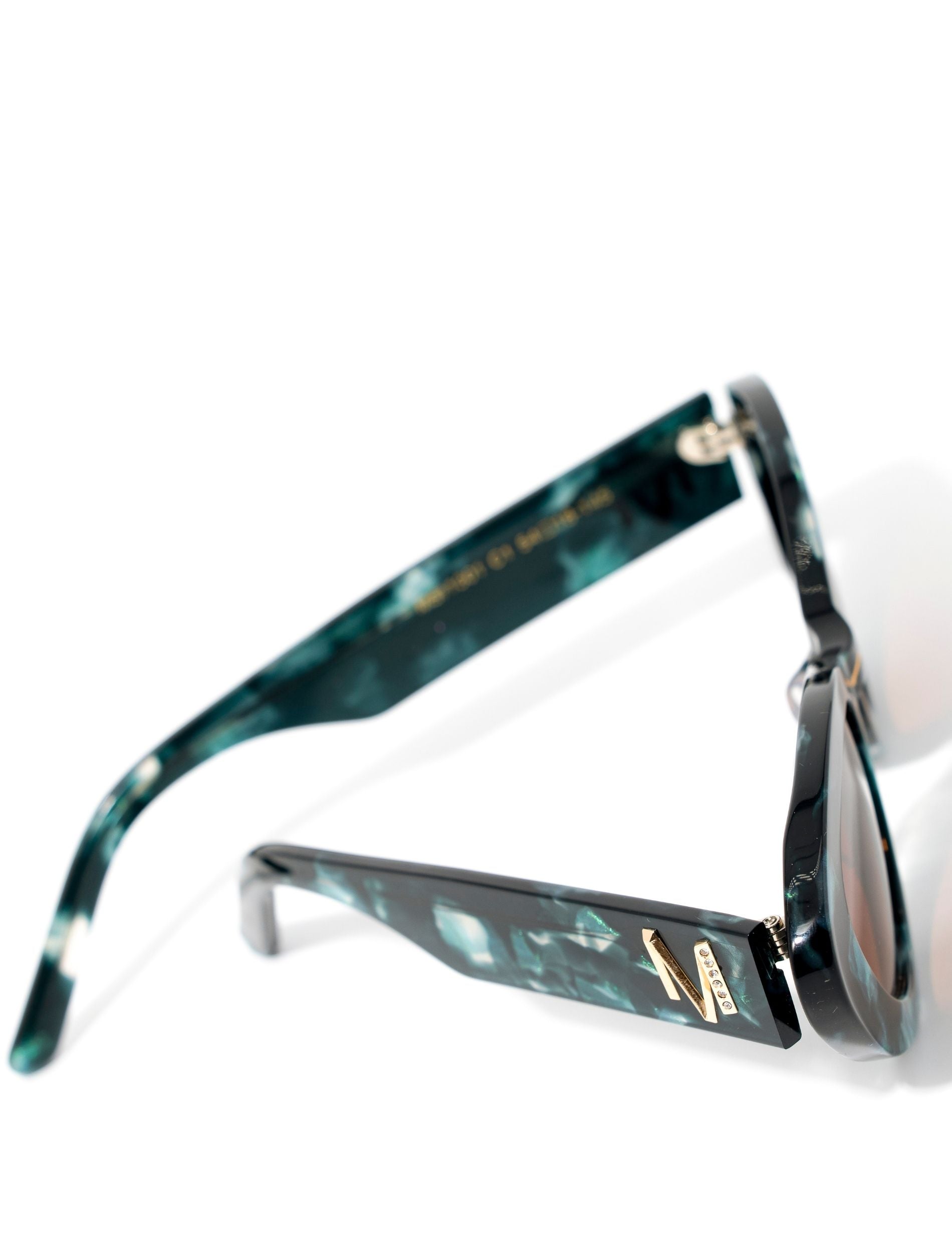 top view of a pair of stylish sunglasses in a green tortoise shell style displaced against a white back drop. the lenses are a gradient brown. You can see an embellished &#39;M&#39; on the side of the frame arm