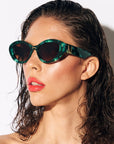 Model with brown hair and glossy red lips, wears a pair of stylish sunglasses with a deep green and black pearlescent frame, set in front white back drop