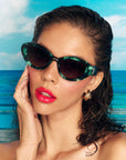 Model with brown hair and glossy red lips, wears a pair of stylish sunglasses with a deep green and black pearlescent frame, set in front of a beach scene and blue sky 