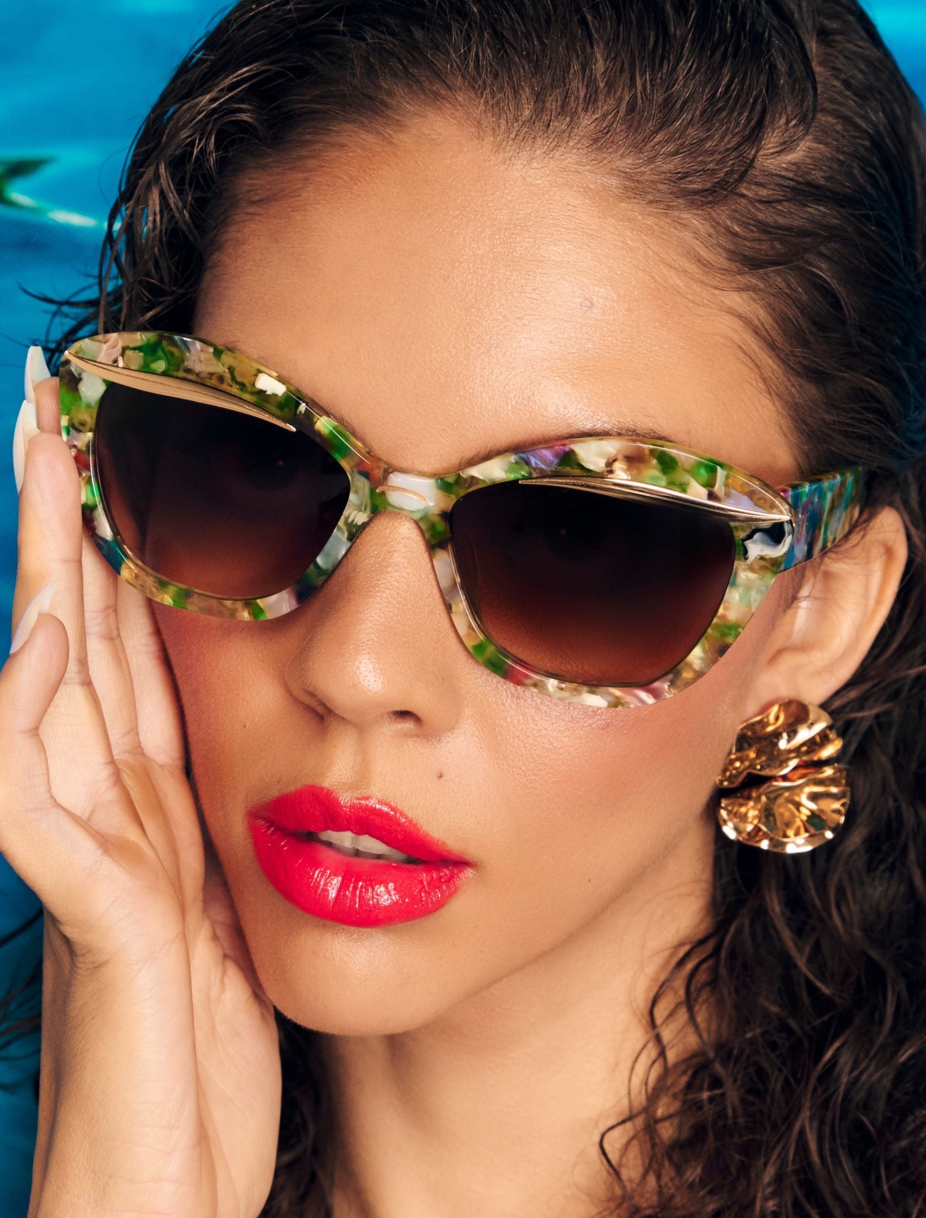 Person wearing colourful green and pink tone sunglasses, golden shell-shaped earrings, and bright red lipstick, holding their cheek against a blue background.