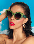 A stylish model with brown hair and glossy red lips wears vibrant green pearlescent sunglasses with gem embellishments on the upper corners on the frame. She is set against a blue sky backdrop