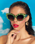 A stylish model with brown hair and glossy red lips wears vibrant green pearlescent sunglasses with gem embellishments on the upper corners on the frame. She is set against a blue sky backdrop