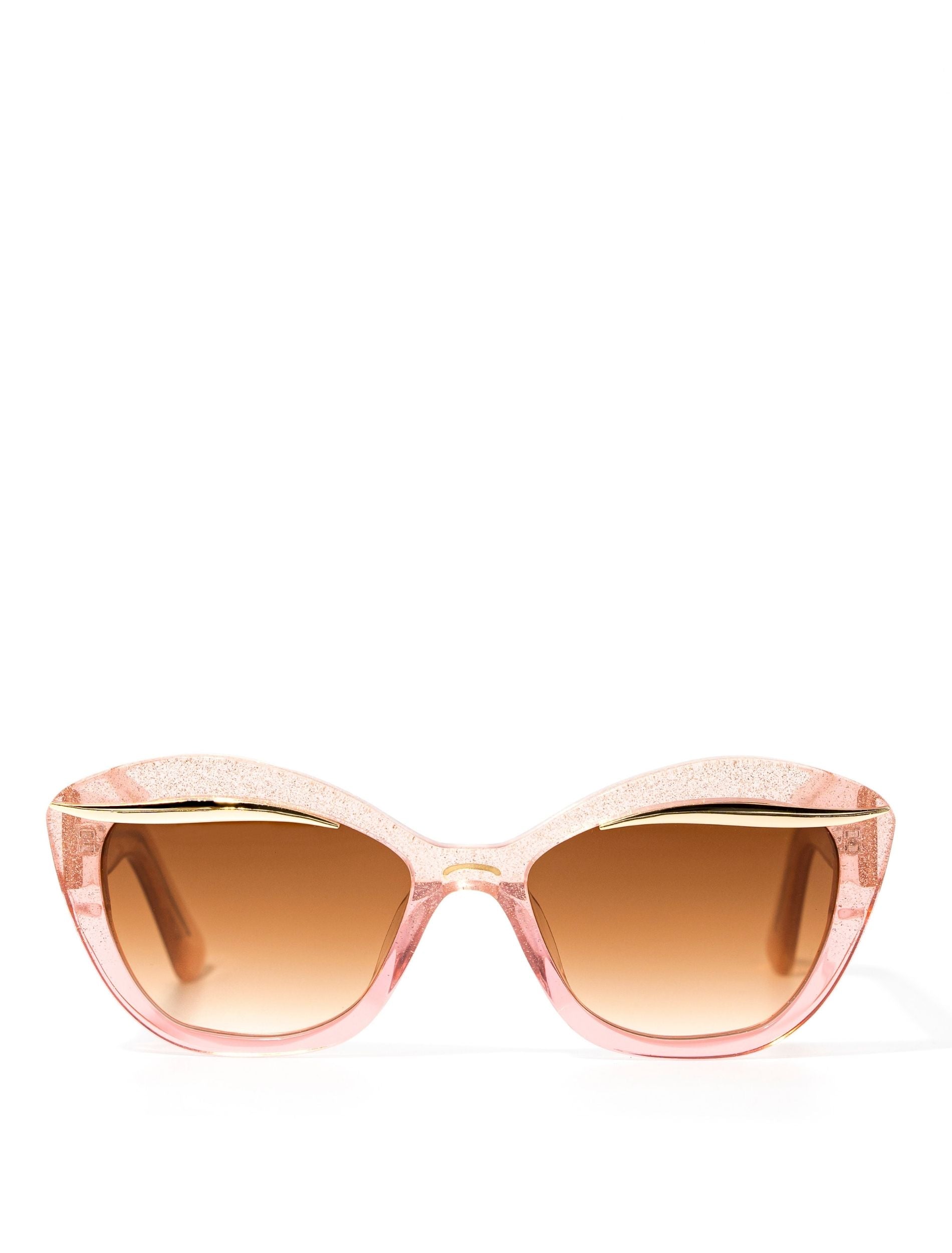 Front view of an elegant pink glittery cat eye framed sunglasses with a gold flick up top of the lenses and a brown gradient lense. Set against a white backdrop