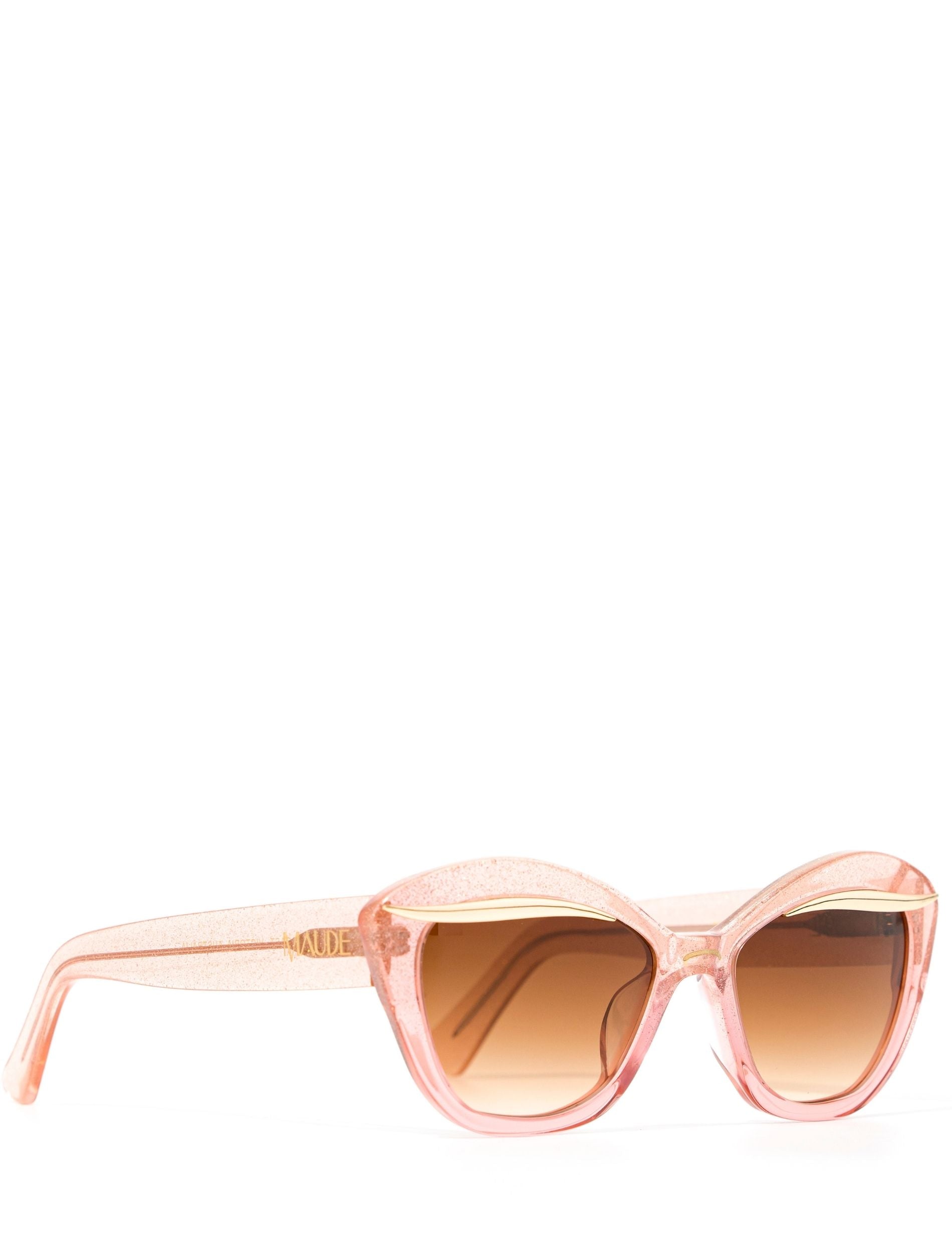 Side view of an elegant pink glittery cat eye framed sunglasses with a gold flick up top of the lenses and a brown gradient lense. Set against a white backdrop. The brand name MAUDE is featured in gold on the arm on the frame