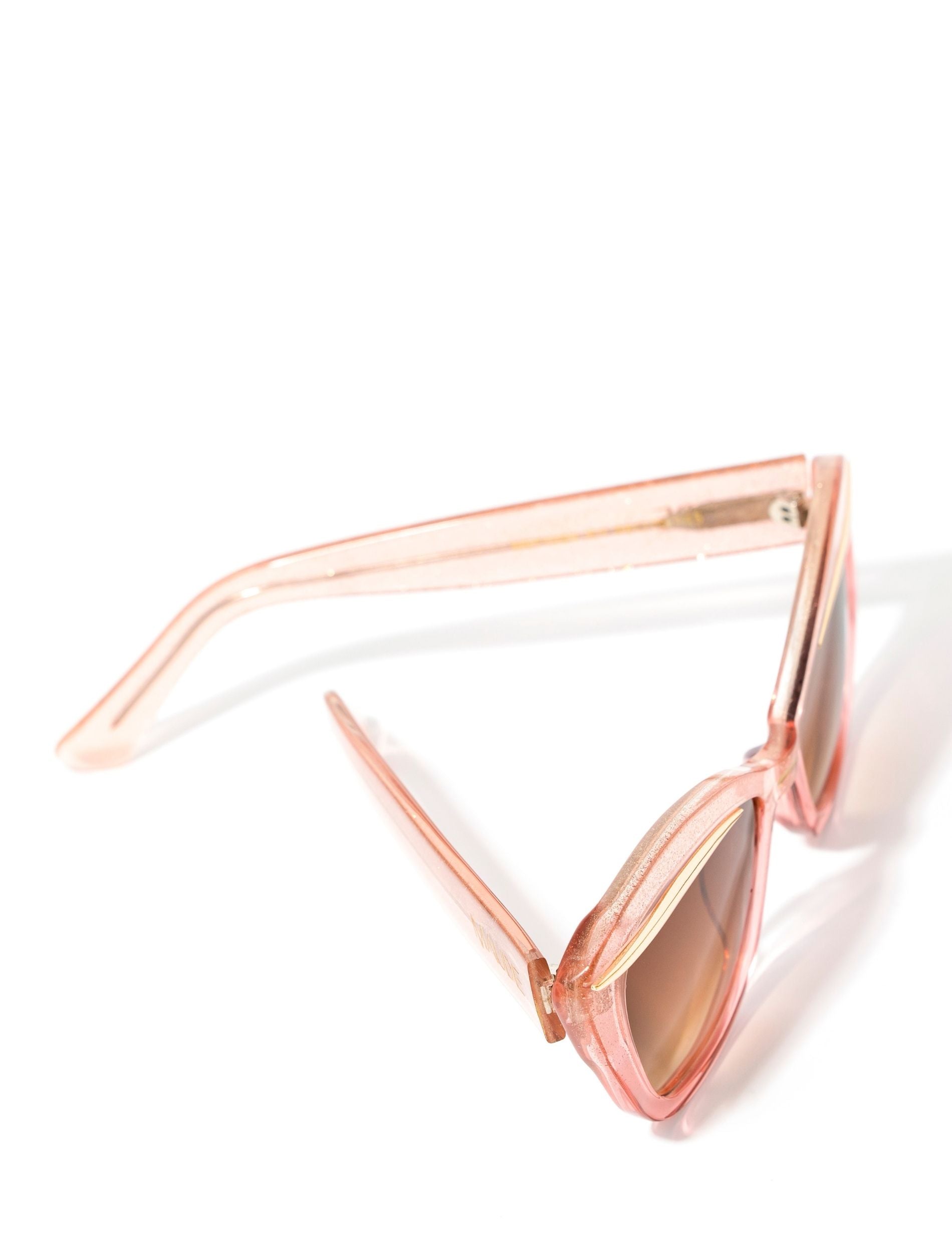 top view of an elegant pink glittery cat eye framed sunglasses with a gold flick featured on top of the lenses. Set against a white backdrop. The brand name MAUDE is featured in gold, subtly on the arm on the frame