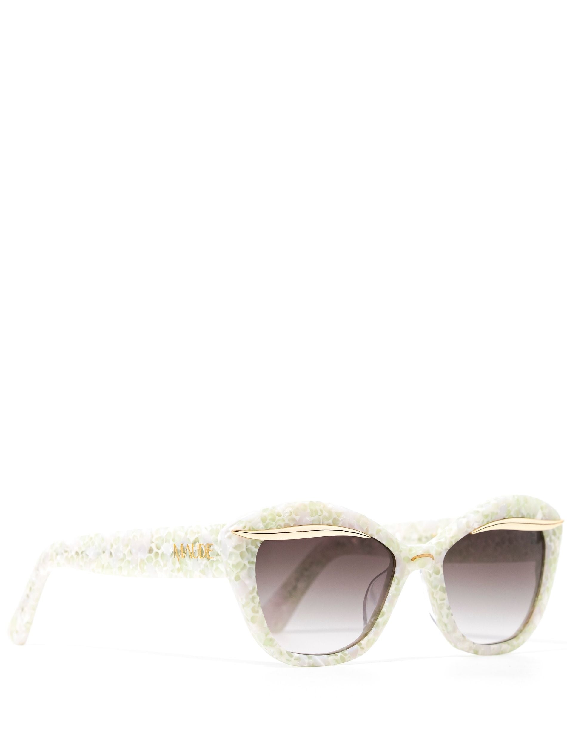 Side view of a stylish pair of sunglasses adorned with a gold flick along the upper lense and a subtle white pearl frame, set against a white backdrop. Featuring a dark grey gradient lense. The words MAUDE are shown on the arm of the sunglasses