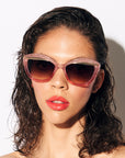 A model with brown hair and glossy red lips wears a stylish pair f sunglasses with a subtle peach pink glitter frame, featuring a gold flick up top of the lenses. the lense is a dark brown gradient. she is set against a white back drop. 