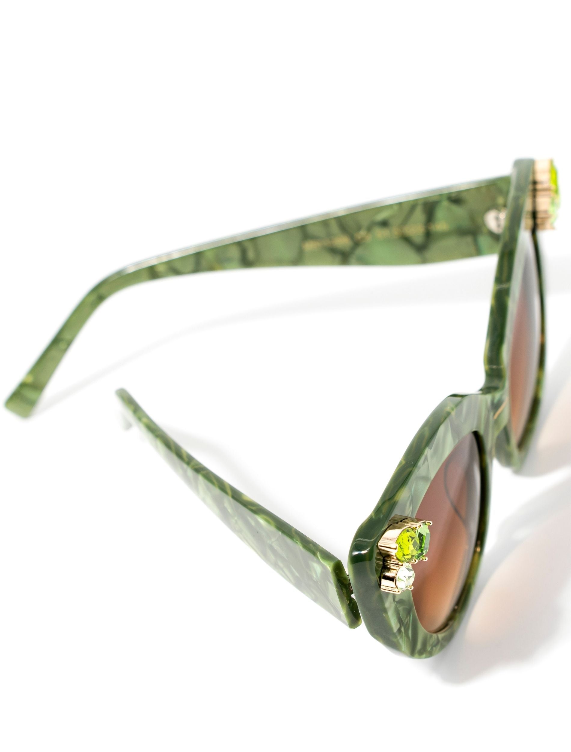 Top view of stunning cat eye sunglasses crafted with a green pearlescent look,  featuring gemstone embellishments on the upper corners of the frame and a brown gradient lense. Set against a white backdrop