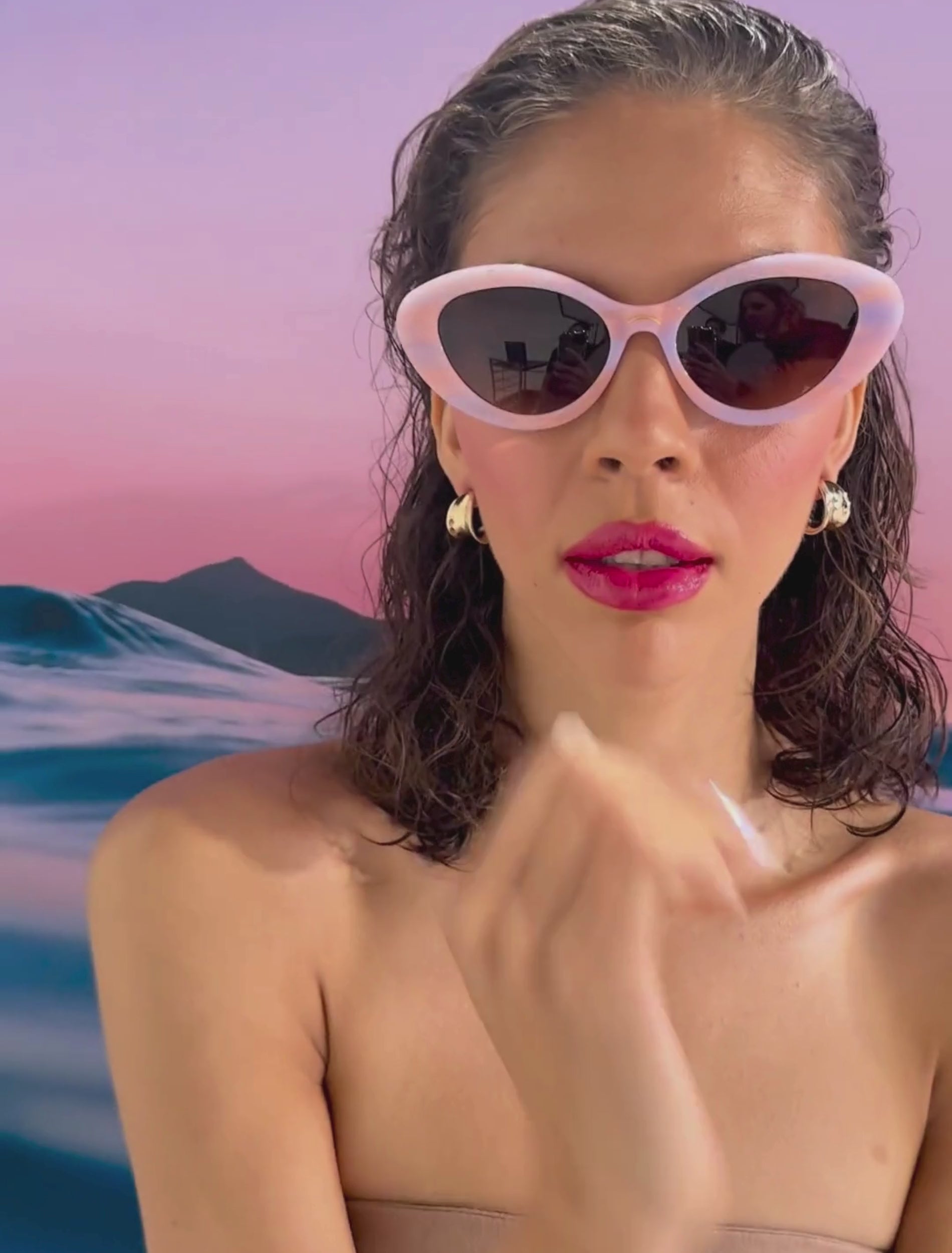 A model with brown hair and glossy red lips wears a pair of pastel pink and purple framed sunglasses in front of a pink sunset background