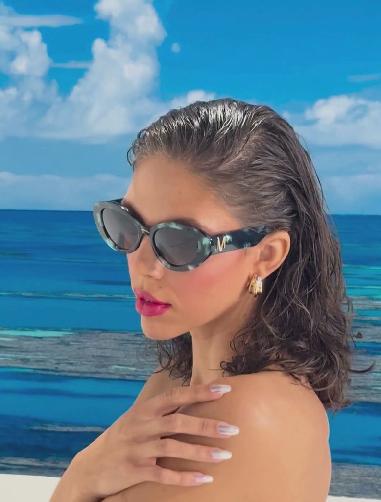 Model with brown hair and glossy red lips, wears a pair of stylish sunglasses with a deep green and black pearlescent frame, set in front of a beach scene and blue sky 