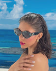 Model with brown hair and glossy red lips, wears a pair of stylish sunglasses with a deep green and black pearlescent frame, set in front of a beach scene and blue sky 