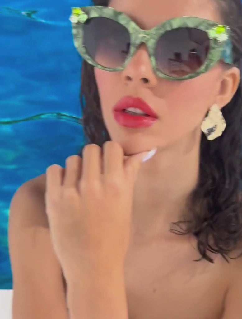 A stylish model with brown hair and glossy red lips wears vibrant green pearlescent sunglasses with gem embellishments on the upper corners on the frame. She is set against a blue ocean backdrop