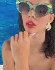 A stylish model with brown hair and glossy red lips wears vibrant green pearlescent sunglasses with gem embellishments on the upper corners on the frame. She is set against a blue ocean backdrop