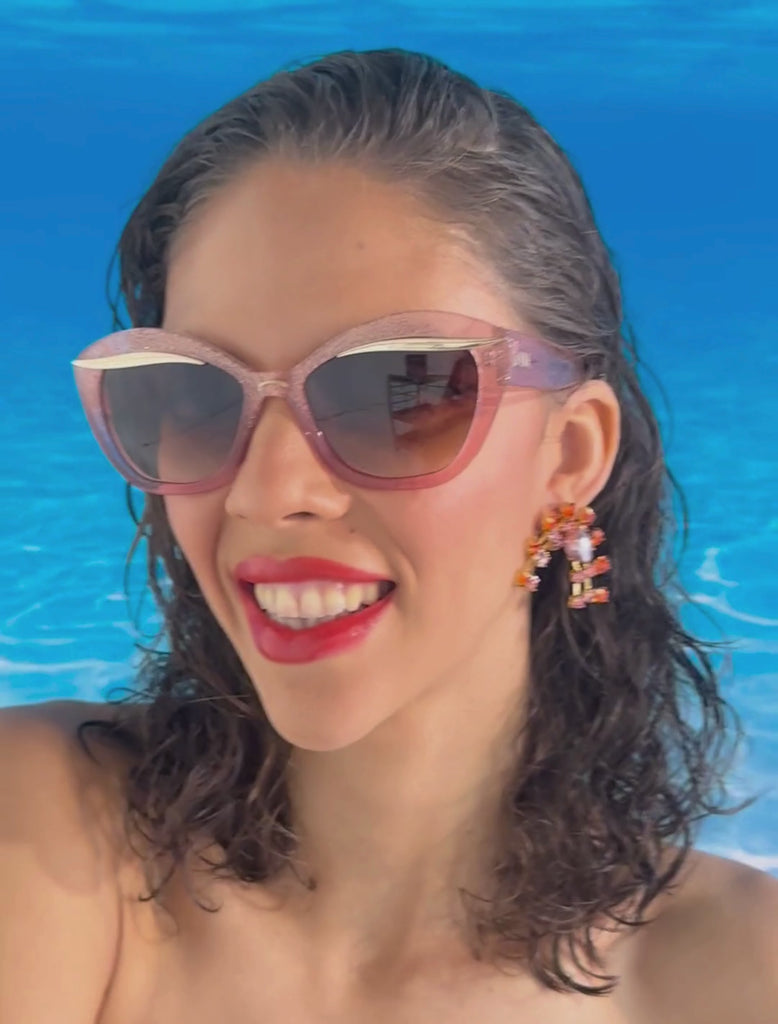 A model with brown hair and glossy red lips wears a stylish pair of sunglasses with a subtle peach pink glitter frame, featuring a gold flick up top of the lenses. the lense is a dark brown gradient. she is set against an ocean blue back drop, she is smiling and showing us the sunglasses