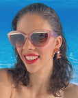 A model with brown hair and glossy red lips wears a stylish pair of sunglasses with a subtle peach pink glitter frame, featuring a gold flick up top of the lenses. the lense is a dark brown gradient. she is set against an ocean blue back drop, she is smiling and showing us the sunglasses